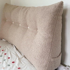 Puede incluir: Una almohada grande, beige y esponjosa con botones capitoné se coloca contra una cabecera blanca en una cama con una colcha blanca con estampado floral.