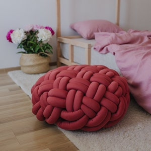 Pode incluir: Uma almofada redonda vermelha trançada está sobre um tapete creme. A almofada é o ponto focal, com uma estrutura de cama e roupa de cama rosa ao fundo. Uma cesta com flores rosa e brancas também é visível.
