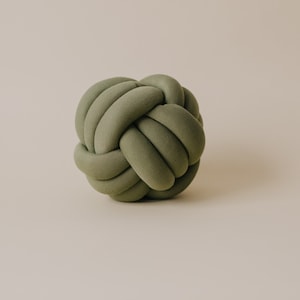 May include: A sage green decorative knot pillow. The pillow is round and made of a soft, textured fabric. The knot design adds a unique and modern touch to home decor. The pillow is a solid color and appears to be plush.