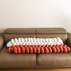 Puede incluir: Un cojín decorativo trenzado en blanco y rojo, colocado sobre un sofá de cuero marrón. El diseño del cojín presenta una serie de segmentos redondeados y conectados. El sofá está frente a una pared neutra, con plantas a ambos lados.