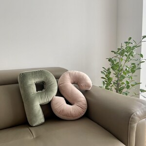 May include: Two plush letter-shaped pillows in a soft velvet fabric. One pillow is shaped like the letter P in a muted sage green color, and the other is shaped like the letter S in a soft blush pink color. Both pillows have a smooth texture.