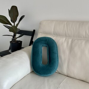 May include: A teal velvet throw pillow shaped like the number zero or the letter O. The pillow features a smooth, soft-touch fabric with a central cutout, resting on a cream-colored leather sofa. The design is simple, rounded, and solid-colored.
