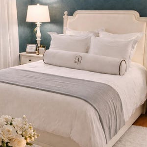 May include: A bedroom with a white bed, white bedding, and a grey throw. Two white bedside tables with lamps either side of the bed. A tufted ottoman with a tray holding flowers, books, and a candle sits in front of the bed.