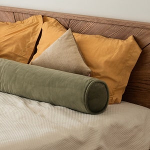 Puede incluir: Un cojín cilíndrico largo de color verde oliva sobre una cama con una colcha texturizada de color crema claro. Detrás hay dos almohadas rectangulares amarillo mostaza y dos cojines triangulares beige contra un cabecero de madera.