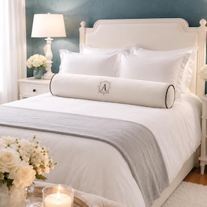 Puede incluir: Un cojín cilíndrico de tela color blanco roto con ribetes oscuros y una letra A bordada en el centro dentro de un marco decorativo. El cojín está colocado horizontalmente sobre una cama blanca con un camino de cama gris y ropa de cama blanca.