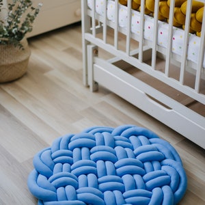 May include: A round, light blue woven rug made of soft fabric. The rug is placed on a light wood floor. A white crib with a yellow braided bumper is in the background, suggesting a nursery setting.