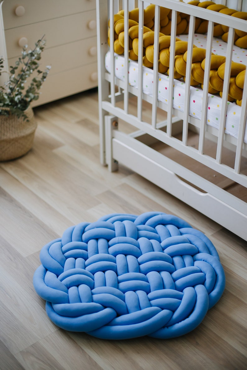 May include: A round, light blue woven rug made of soft fabric. The rug is placed on a light wood floor. A white crib with a yellow braided bumper is in the background, suggesting a nursery setting.