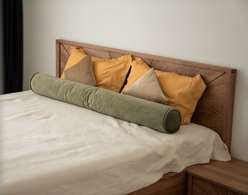 May include: A bed with a wooden headboard and a cream-colored blanket. The bed is adorned with mustard-yellow and tan pillows, and a long, cylindrical, olive-green bolster pillow. The headboard and nightstand are made of wood.