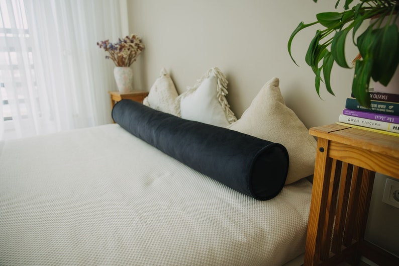 May include: A long, cylindrical bolster pillow in a deep navy blue velvet fabric rests on a white bedspread. The pillow is approximately 30 inches long and is a decorative element for a bed or sofa.