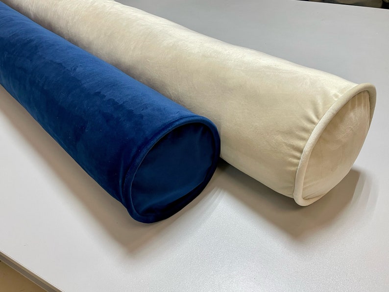 May include: Two cylindrical bolster pillows. One is a deep navy blue, the other is a soft cream color. Both are made of a plush, velvety fabric. The ends of the pillows are rounded and finished with a contrasting trim.