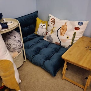 May include: A dark blue tufted floor cushion with a backrest, accompanied by a small yellow square pillow featuring a white cat illustration and a large rectangular white pillow decorated with colorful abstract shapes, vases, and botanical patterns.