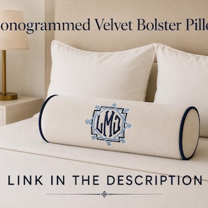 May include: A cream-colored velvet bolster pillow featuring dark blue piping on the ends and a central embroidered monogram with the letters LMG inside a geometric frame. The text Monogrammed Velvet Bolster Pillow and Link in the description appear.