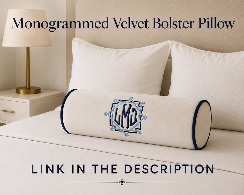 May include: A cream-colored velvet bolster pillow featuring dark blue piping on the ends and a central embroidered monogram with the letters LMG inside a geometric frame. The text Monogrammed Velvet Bolster Pillow and Link in the description appear.