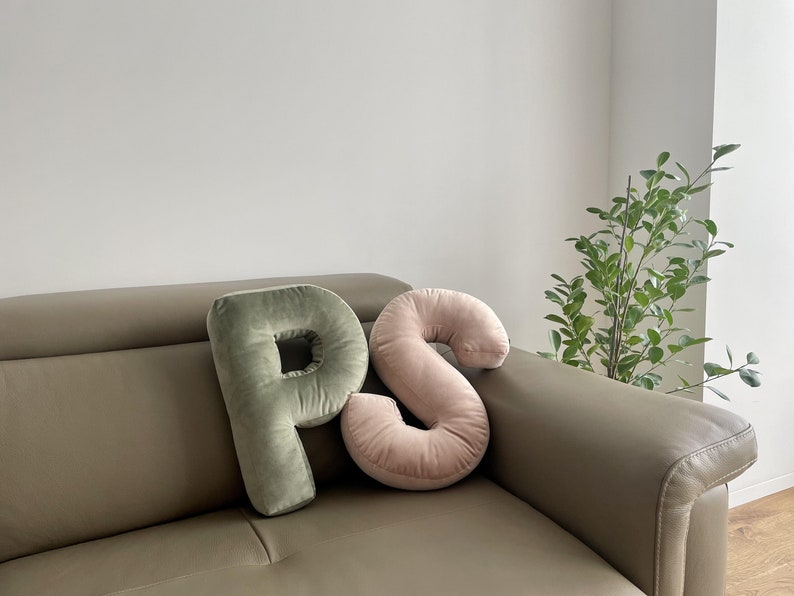 May include: Two plush letter-shaped pillows in a soft velvet fabric. One pillow is shaped like the letter P in a muted sage green color, and the other is shaped like the letter S in a soft blush pink color. Both pillows have a smooth texture.