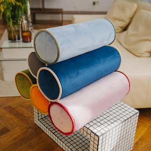 Könnte beinhalten: Ein Stapel aus fünf zylindrischen Kissen in verschiedenen Farben, darunter Blau, Rosa, Orange, Grün und Grau. Die Kissen sind auf einem weißen und schwarzen gekachelten Würfel gestapelt.