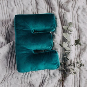 May include: A plush, deep teal velvet pillow shaped like the capital letter E. The pillow has a smooth, soft texture and is positioned next to a single, slender stem with small, round, muted green leaves, resting on a light gray cable knit fabric.