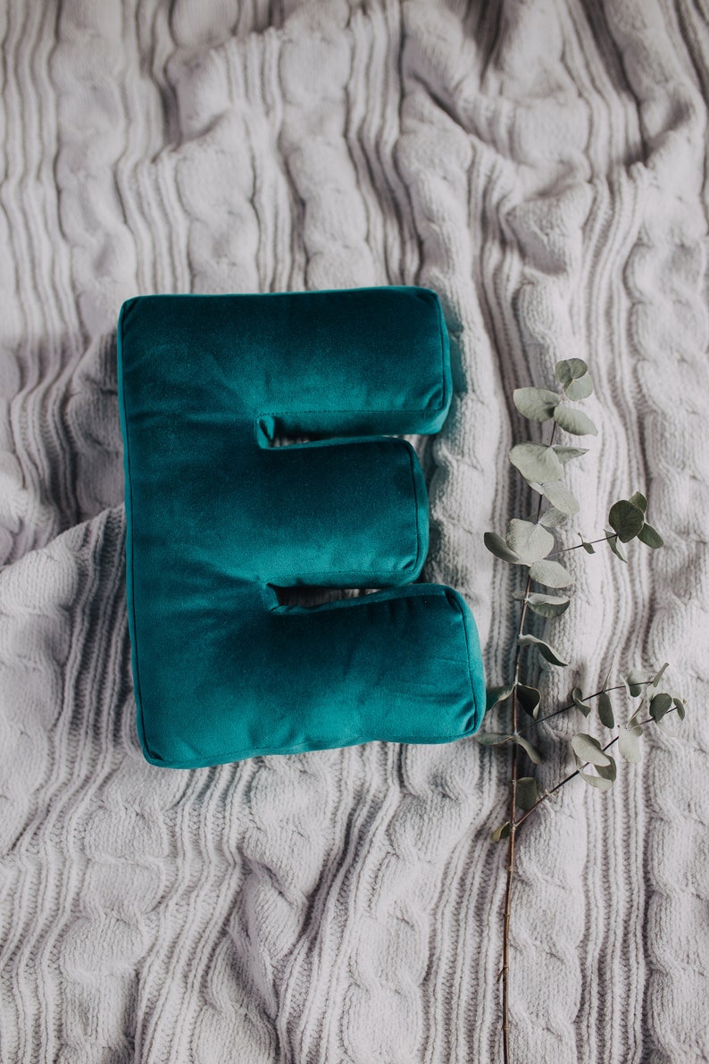 May include: A plush, deep teal velvet pillow shaped like the capital letter E. The pillow has a smooth, soft texture and is positioned next to a single, slender stem with small, round, muted green leaves, resting on a light gray cable knit fabric.