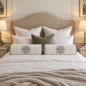 Op de afbeelding: Twee cilindervormige bolsterkussens van gebroken wit textiel met een donkergrijs geborduurd monogram met de letters MOR in een geometrisch kader. De kussens liggen horizontaal op een bed met witte en olijfgroene sierkussens.