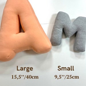Może przedstawiać: Dwie litery z tkaniny, pomarańczowa "A" i szara "M". Litera "A" jest oznaczona jako "Large" i mierzy 40 cm. Litera "M" jest oznaczona jako "Small" i mierzy 25 cm. Litery są wyświetlane na białym tle.