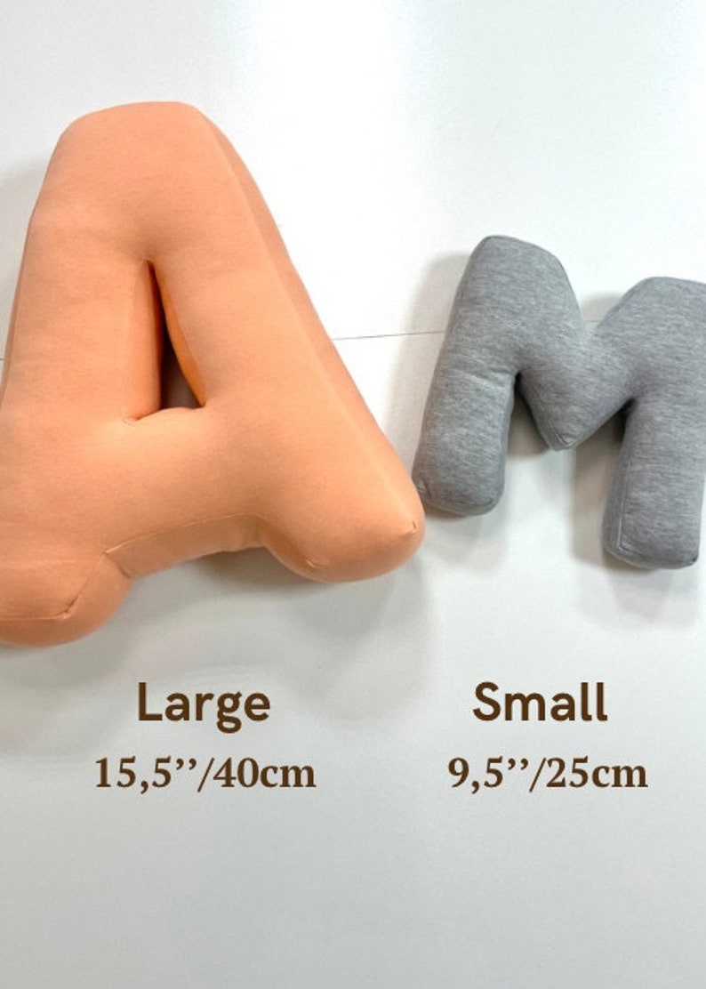 Może przedstawiać: Dwie litery z tkaniny, pomarańczowa "A" i szara "M". Litera "A" jest oznaczona jako "Large" i mierzy 40 cm. Litera "M" jest oznaczona jako "Small" i mierzy 25 cm. Litery są wyświetlane na białym tle.