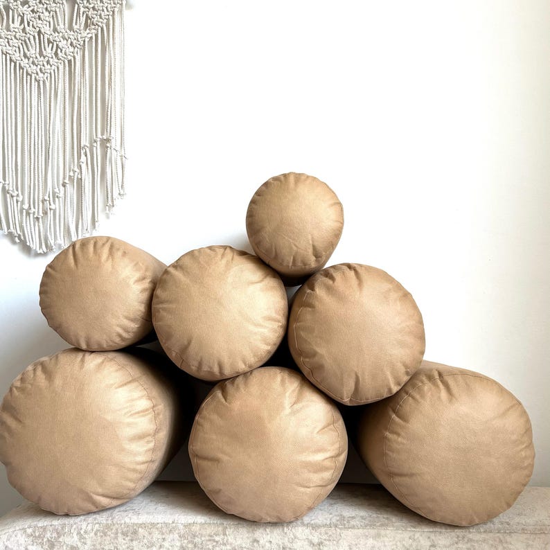 May include: A stack of six brown cylindrical pillows arranged in a pyramid shape. The pillows are made of a soft, textured fabric.