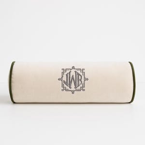 May include: A cylindrical bolster pillow in a light cream color with dark olive green piping along the edges. The center features an embroidered monogram with the letters JWB enclosed within a geometric, square-shaped decorative frame.