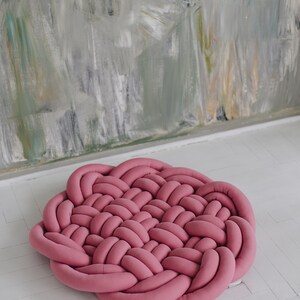 May include: A round, braided rug in a dusty rose color. The rug is made of soft, woven material and sits on a white floor. The background features a textured wall with muted colors.