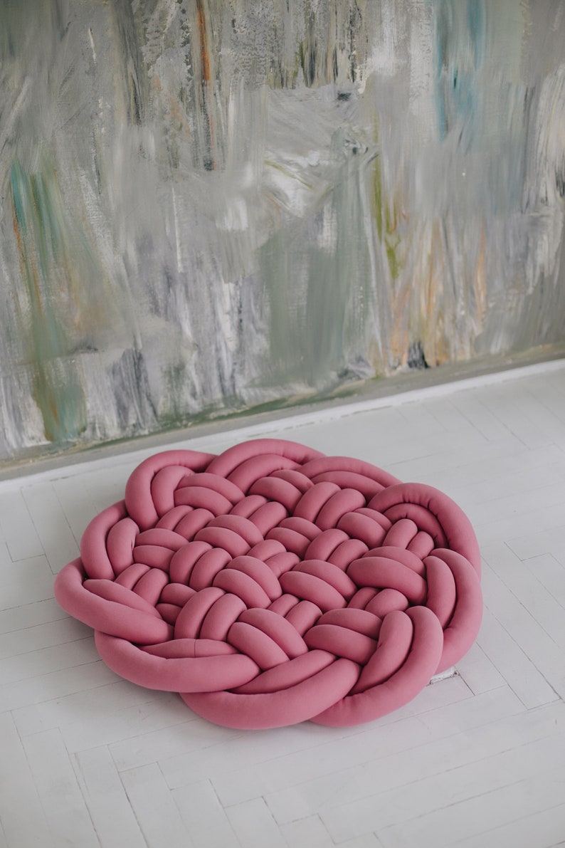 May include: A round, braided rug in a dusty rose color. The rug is made of soft, woven material and sits on a white floor. The background features a textured wall with muted colors.