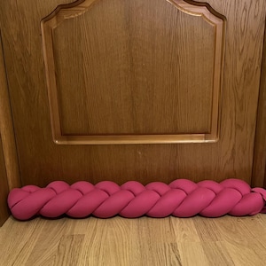 May include: A pink braided doorstop made of fabric. The doorstop is placed at the bottom of a wooden door.