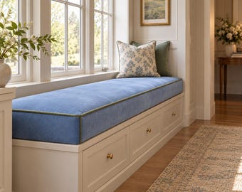 May include: A white built-in window bench featuring a long blue upholstered cushion with green piping. The base includes three storage drawers with small round brass knobs. Two decorative throw pillows are placed on the bench.