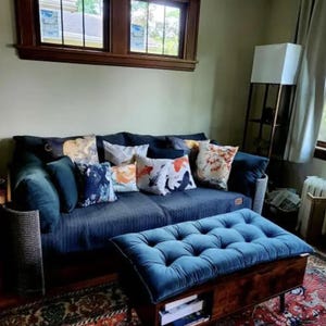 May include: A dark blue sofa with a tufted blue ottoman in front. The sofa features several decorative throw pillows with various patterns, including koi fish and floral designs. The furniture is set against a wall with a large wooden window frame.