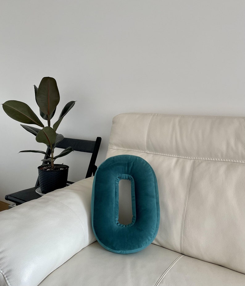 May include: A teal velvet throw pillow shaped like the number zero or the letter O. The pillow features a smooth, soft-touch fabric with a central cutout, resting on a cream-colored leather sofa. The design is simple, rounded, and solid-colored.