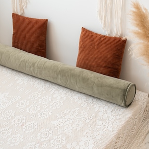 May include: A long, cylindrical sage green velvet bolster pillow positioned horizontally across a bed covered with a white lace bedspread, accompanied by two square, rust-colored corduroy throw pillows placed against the wall.