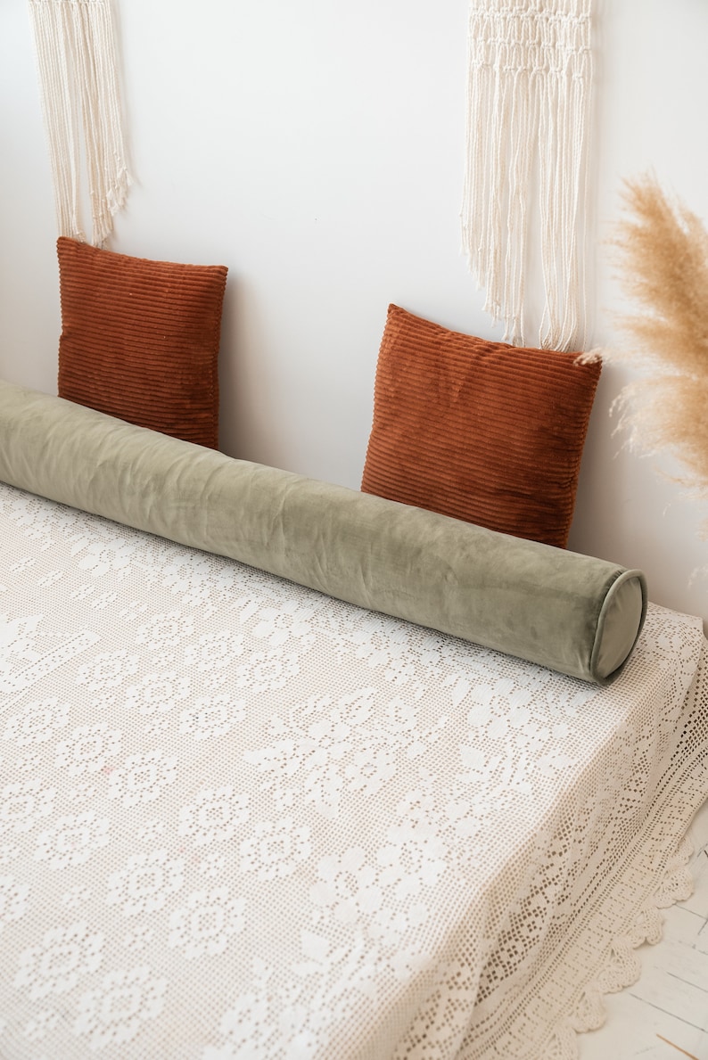 May include: A long, cylindrical sage green velvet bolster pillow positioned horizontally across a bed covered with a white lace bedspread, accompanied by two square, rust-colored corduroy throw pillows placed against the wall.