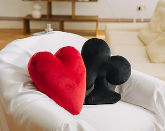 May include: Two plush pillows shaped like a heart and a club, in red and black velvet, respectively, are sitting on a white armchair.