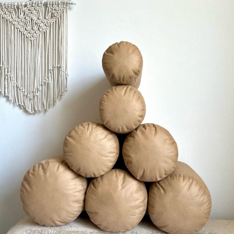 May include: A stack of seven brown cylindrical pillows arranged in a triangular shape. The pillows are made of a soft, textured fabric.