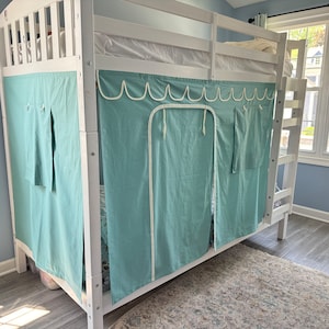May include: A white bunk bed with a teal blue fabric canopy. The canopy has a white trim and a pocket on each side. The bed is in a room with a window and a rug.