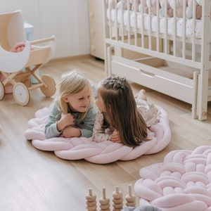 May include: Two young girls are lying on a large, pink braided floor mat. The mat is in a room with a crib, a doll carriage, and other children's toys. The floor is made of wood.