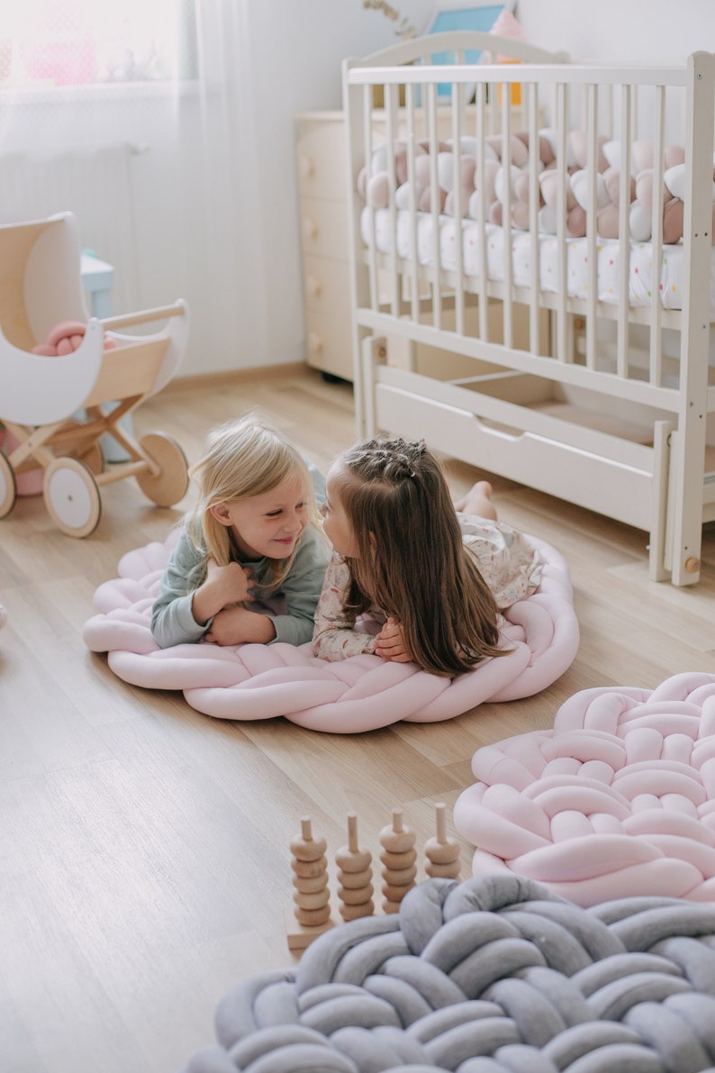 May include: Two young girls are lying on a large, pink braided floor mat. The mat is in a room with a crib, a doll carriage, and other children's toys. The floor is made of wood.