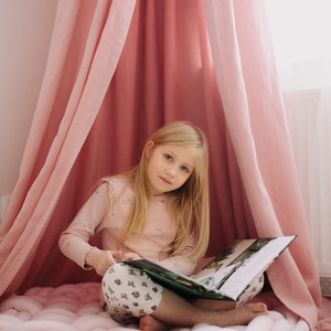 May include: A young girl with long blonde hair sits inside a pink canopy, reading a book. She wears a light pink long-sleeved shirt and patterned pants. A large, braided pink floor cushion is beneath her.