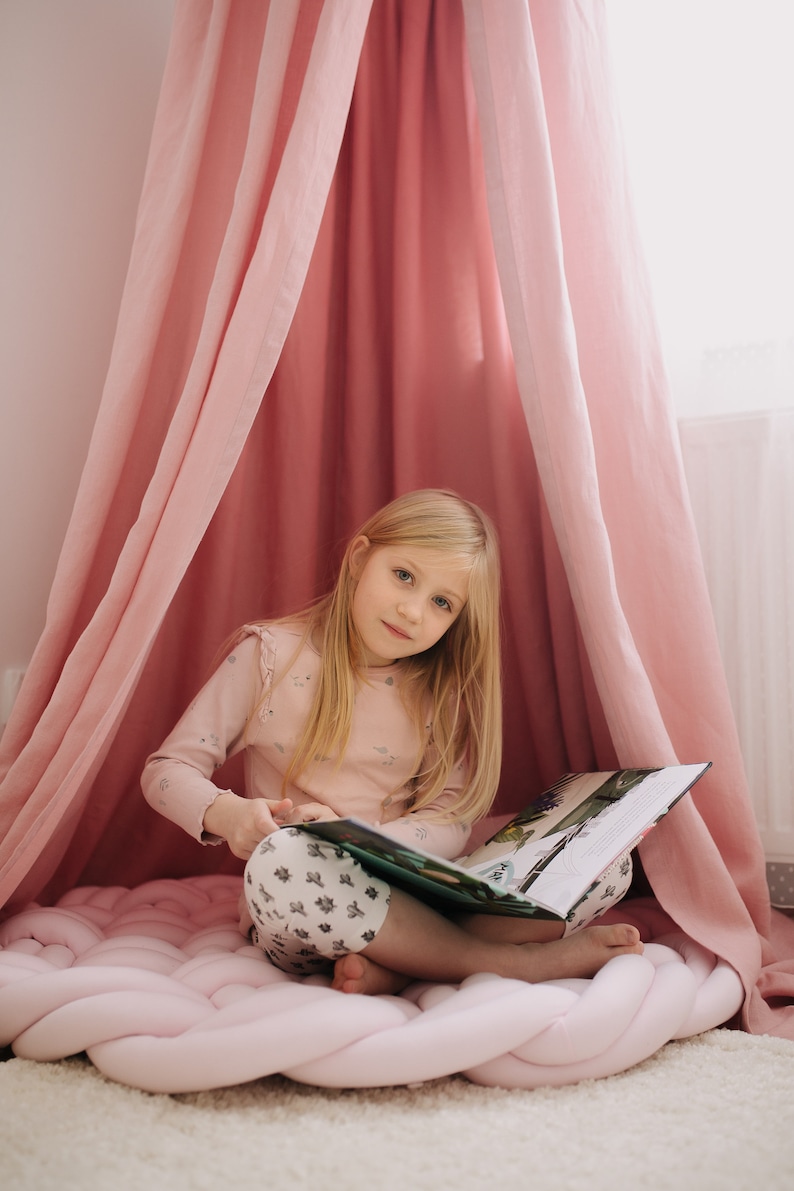 May include: A young girl with long blonde hair sits inside a pink canopy, reading a book. She wears a light pink long-sleeved shirt and patterned pants. A large, braided pink floor cushion is beneath her.