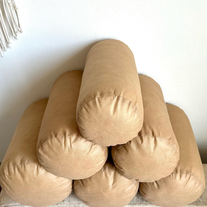 May include: A stack of six brown cylindrical pillows arranged in a pyramid shape. The pillows are made of a soft, textured fabric.