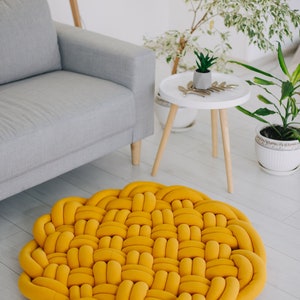 May include: A round, woven rug in a vibrant yellow hue. The rug's design features a repeating pattern of interwoven, tube-like elements. It's placed on a light-colored wooden floor, near a gray sofa and potted plants, creating a cozy interior design.