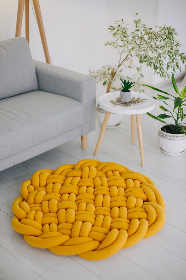 May include: A round, woven rug in a vibrant yellow hue. The rug's design features a repeating pattern of interwoven, tube-like elements. It's placed on a light-colored wooden floor, near a gray sofa and potted plants, creating a cozy interior design.