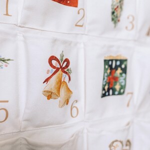 May include: White fabric advent calendar with numbered pockets. Each pocket features a watercolor illustration of Christmas-themed images, including bells, ornaments, and gifts. The numbers are in a gold color.