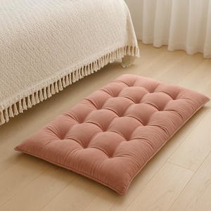 May include: A rectangular floor cushion in a dusty rose color featuring a tufted design with multiple fabric-covered buttons across the surface. The cushion has a soft, plush texture and is positioned flat on a light-colored wooden floor surface.