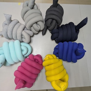 Op de afbeelding: Een bovenaanzicht toont een verzameling geknoopte stoffen decoratieve kussens in verschillende kleuren. De kleuren zijn lichtgrijs, donkergrijs, marineblauw, blauw, geel, roze en teal. De kussens zijn in een cirkel gerangschikt.