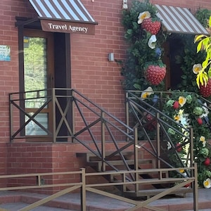 Puede incluir: Un edificio de ladrillo rojo con una entrada de agencia de viajes con un toldo a rayas marrones y blancas. Una barandilla de metal rodea los escalones y una decoraci&oacute;n de pared incluye fresas artificiales grandes y follaje verde.