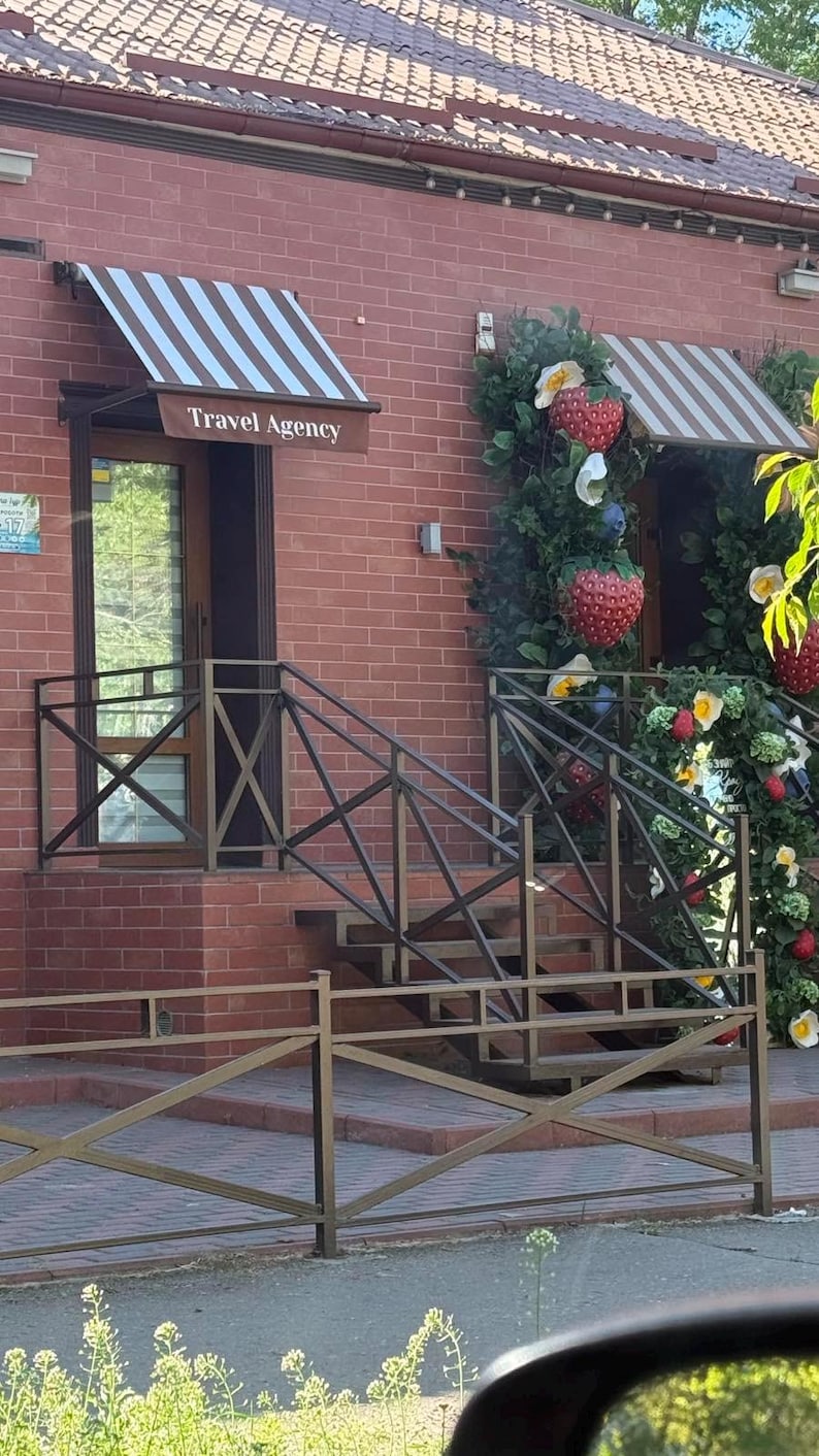 Puede incluir: Un edificio de ladrillo rojo con una entrada de agencia de viajes con un toldo a rayas marrones y blancas. Una barandilla de metal rodea los escalones y una decoraci&oacute;n de pared incluye fresas artificiales grandes y follaje verde.