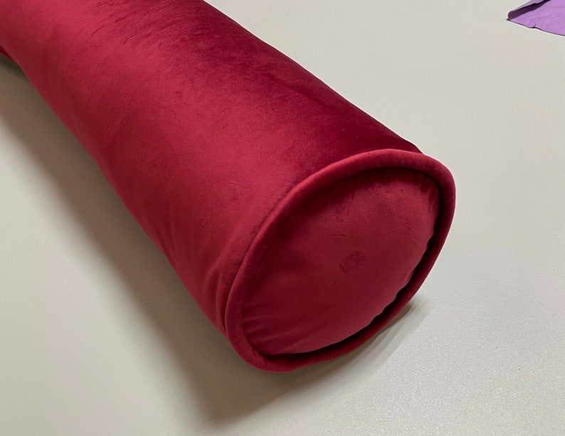May include: A burgundy cylindrical bolster pillow made of soft velvet fabric. The pillow has rounded ends and a smooth, plush texture. The color is a deep, rich red, adding a touch of elegance to any space.